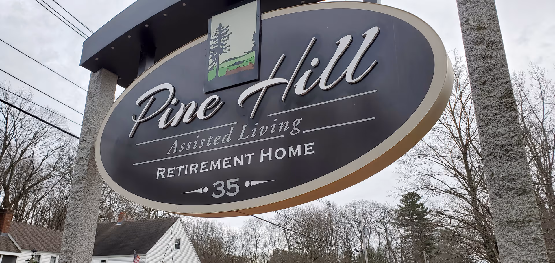 Large oval sign for Pine Hill Assisted Living Retirement Home with the number 35, mounted on two stone pillars with trees and houses in the background under a cloudy sky.