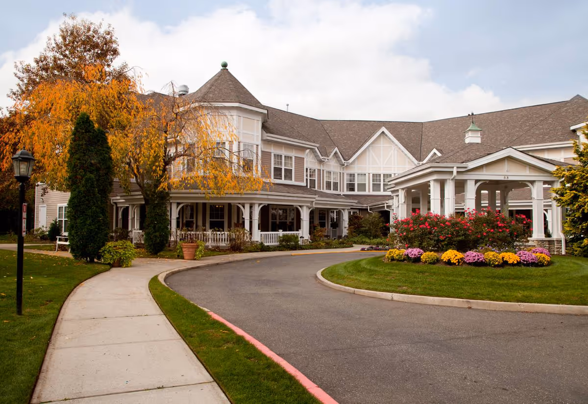 Front entrance of a two-story senior living building with a curved driveway, covered portico, and landscaped flowerbeds.
