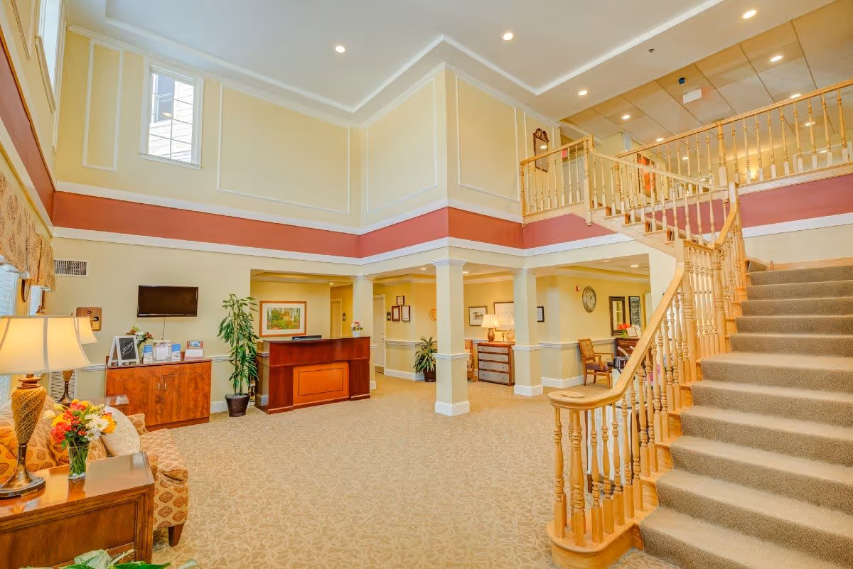 A bright and spacious interior lobby area with a beige carpet and light yellow walls accented by a red stripe. There is a wooden staircase with carpeted steps leading to an upper floor. The room features a reception desk, a small seating area with a patterned armchair, a side table with a lamp and flowers, and various plants and framed artwork on the walls. The ceiling has recessed lighting and crown molding.