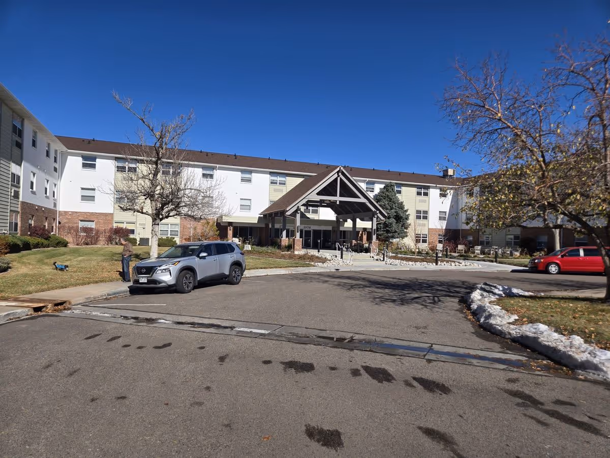 Front entrance of a three-story senior living building with a covered porte-cochère, parked cars, lawn and trees under a clear blue sky.