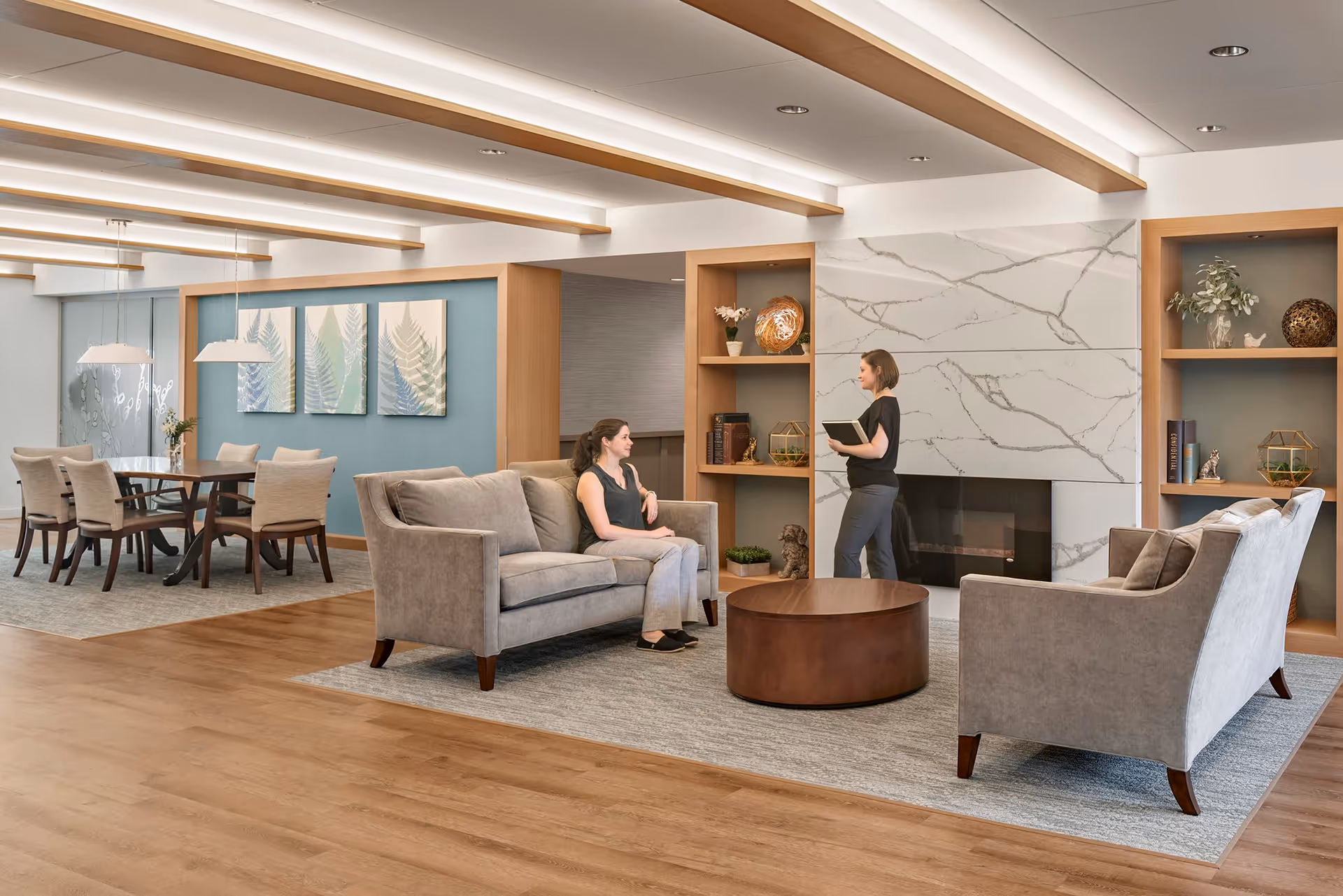 A modern living room area in a senior living facility with two women interacting. One woman is sitting on a gray sofa while the other stands holding a book near a fireplace with marble-like wall panels. The room features wooden shelves with decorative items, a round wooden coffee table, and a dining area with a table and chairs in the background. The space has wooden flooring and ceiling beams with recessed lighting.