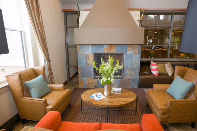 A cozy living room area with two brown leather armchairs with blue cushions, a red sofa, a round wooden coffee table with a vase of white and green flowers, and a fireplace with a stone surround. There are large windows with curtains on the left and a brown leather bench with colorful pillows in the background.