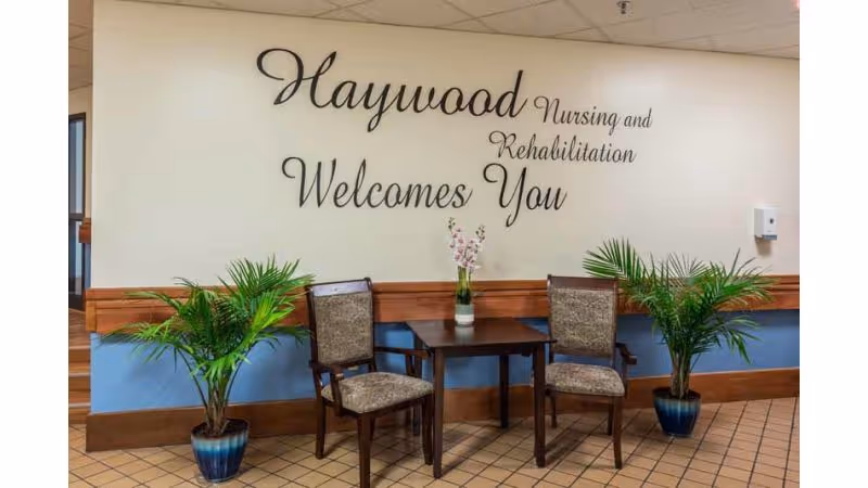 A small seating area with two upholstered chairs and a wooden table between them, holding a vase with flowers. Two potted plants flank the seating area. On the wall behind, decorative text reads 'Haywood Nursing and Rehabilitation Welcomes You'. The floor has a tiled pattern and the lower part of the wall is painted blue with wooden trim.