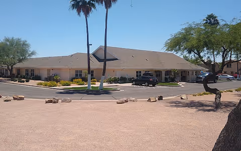 Single-story building with a beige exterior and a brown roof, surrounded by a paved driveway and desert landscaping including palm trees and small bushes under a clear blue sky.