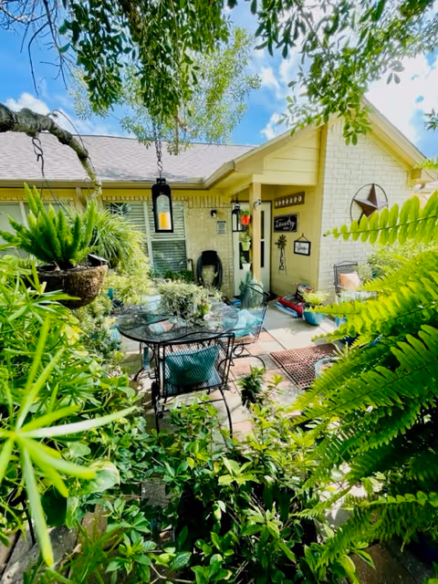 A lush outdoor patio area at La Vista Retirement Community featuring a glass-top table with metal chairs surrounded by abundant green plants and ferns. The patio is adjacent to a light yellow brick building with a star decoration on the wall and various hanging lanterns and signs. The sky is blue with some clouds visible through the tree branches overhead.