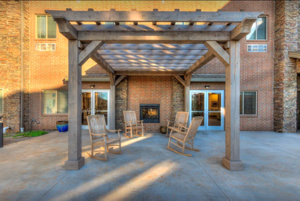 Outdoor patio area at Teal Creek Senior Living featuring a wooden pergola with four wooden rocking chairs arranged around a built-in brick fireplace. The patio is paved with concrete and the building exterior is made of brick with several windows and glass doors.