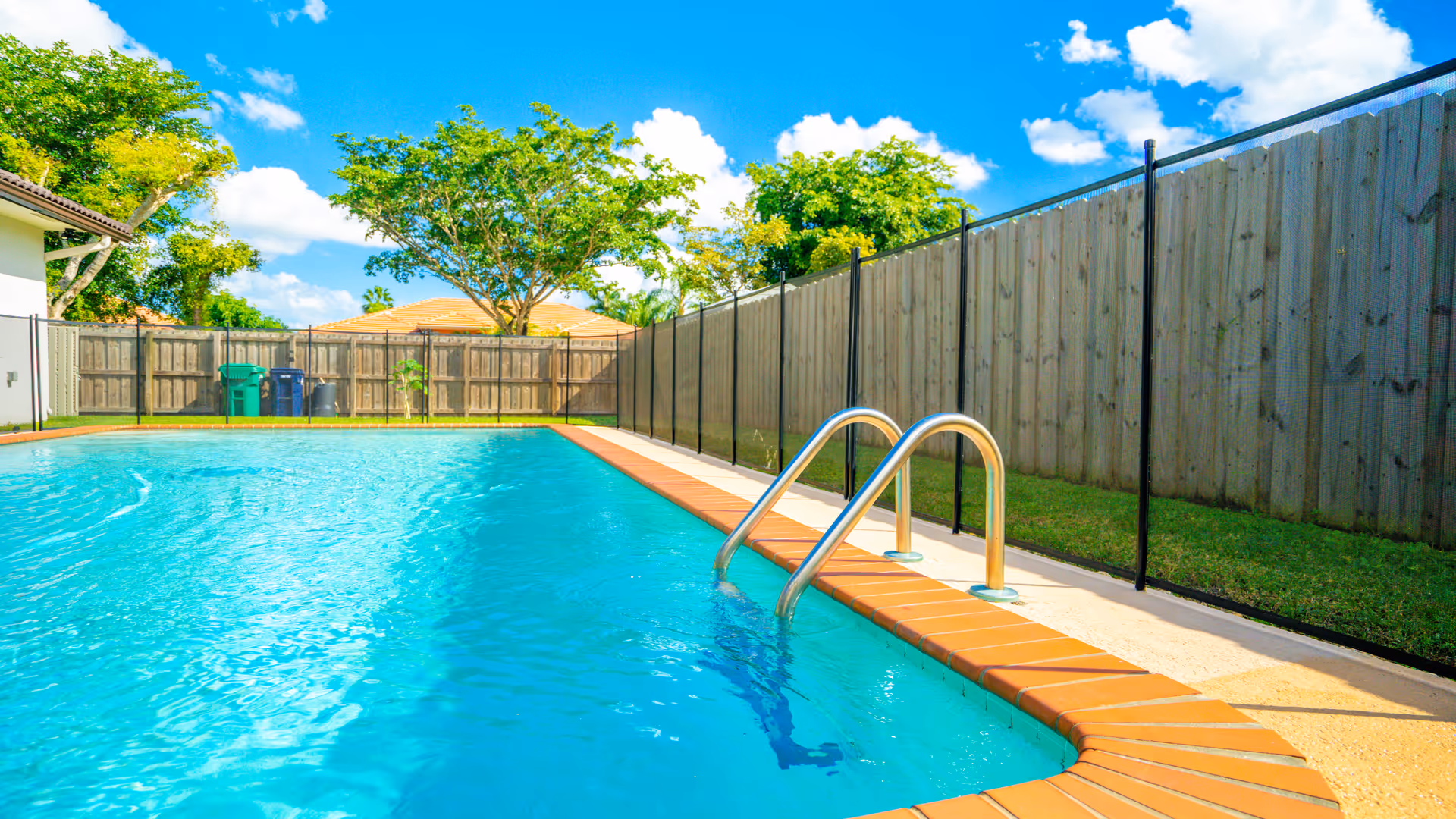 Outdoor swimming pool with clear blue water, surrounded by a brick edge and a safety fence. There are trees and a wooden fence in the background under a bright blue sky with some clouds.