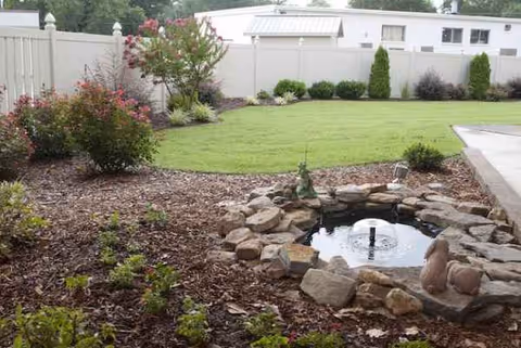 Outdoor landscape with a stone fountain, trimmed lawn, shrubs, and a white fence.
