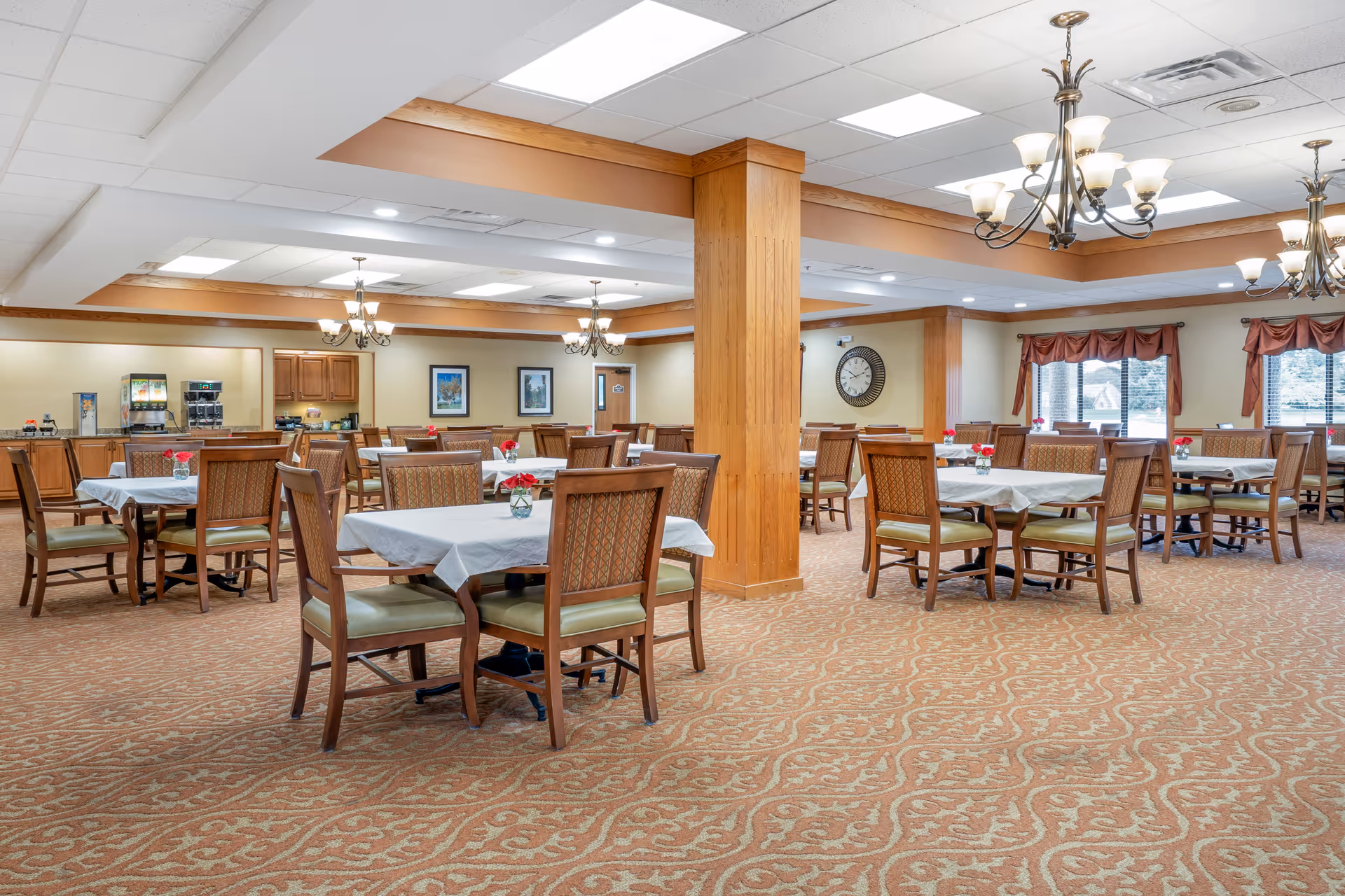 A spacious dining room with multiple tables covered with white tablecloths and surrounded by wooden chairs with green cushions. The room features patterned carpet, wooden columns, chandeliers, large windows with red valances, and a beverage station in the background.