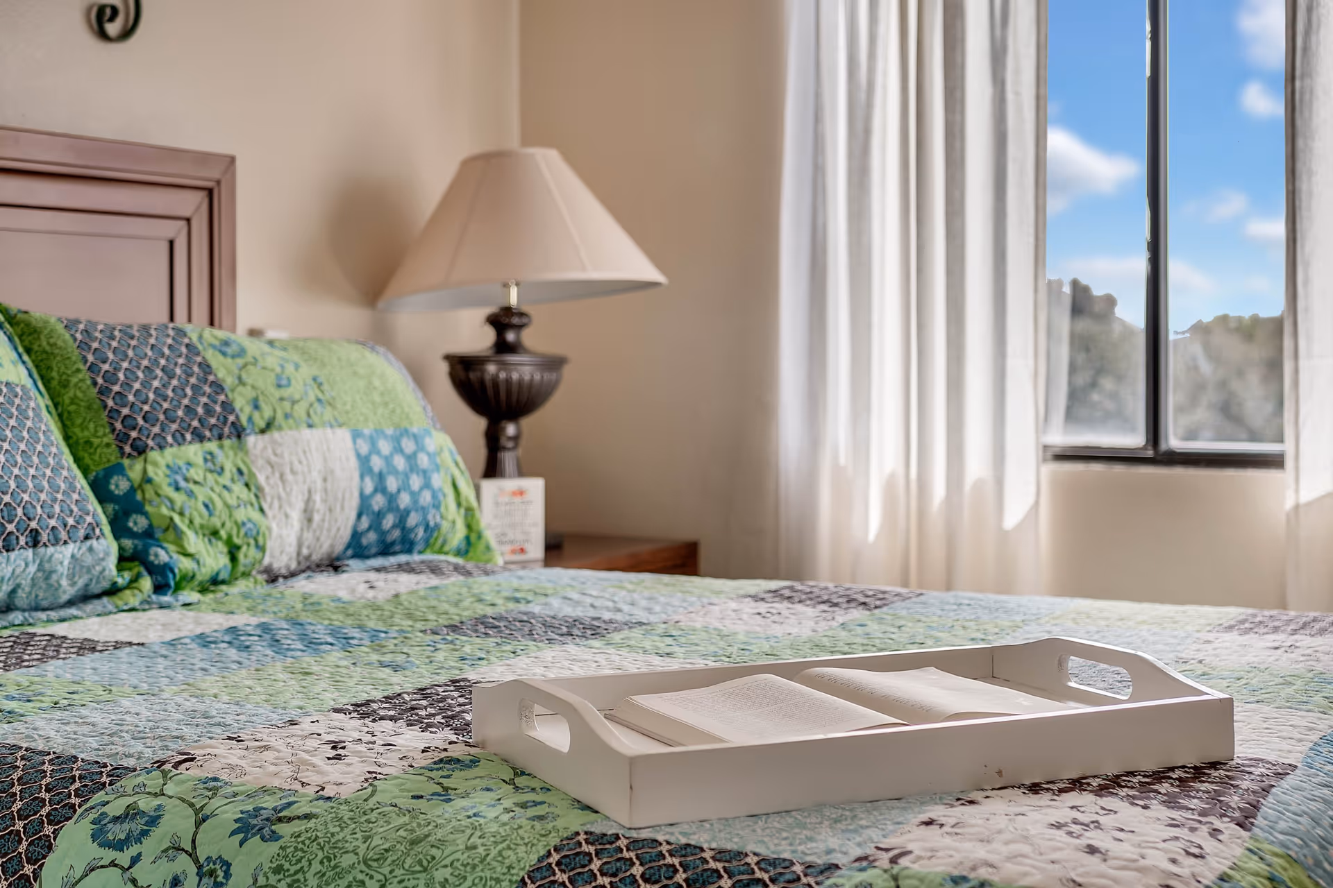 A cozy bedroom with a bed covered in a green, blue, and white patchwork quilt. On the bed is a white tray holding an open book. A bedside table with a dark lamp and a window with sheer white curtains letting in natural light are visible in the background.