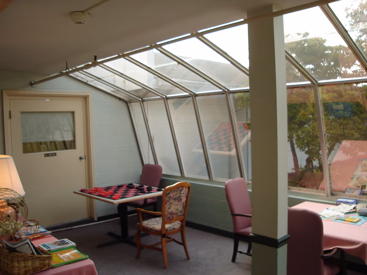 A sunlit indoor seating area with large slanted windows showing an outdoor view with trees. The room contains a table with a checkerboard game and two chairs, another table with magazines and two more chairs, a lamp, and a basket with books or magazines. The walls are light-colored, and there is a door labeled 'No Smoking'.
