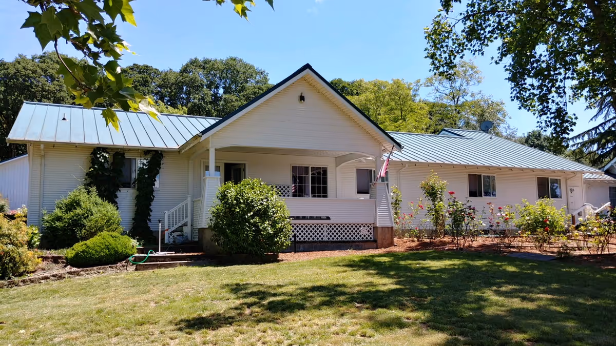 A single-story white building with a metal roof surrounded by green trees and bushes under a clear blue sky. The building has a small porch with steps leading up to it and a garden area with flowers in front.