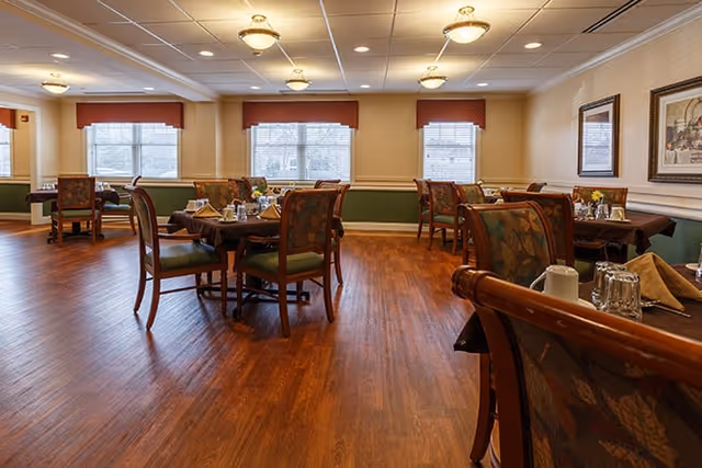 A spacious dining room with wooden floors, several tables set with tableware, chairs with patterned upholstery, large windows with blinds, and framed artwork on the walls.