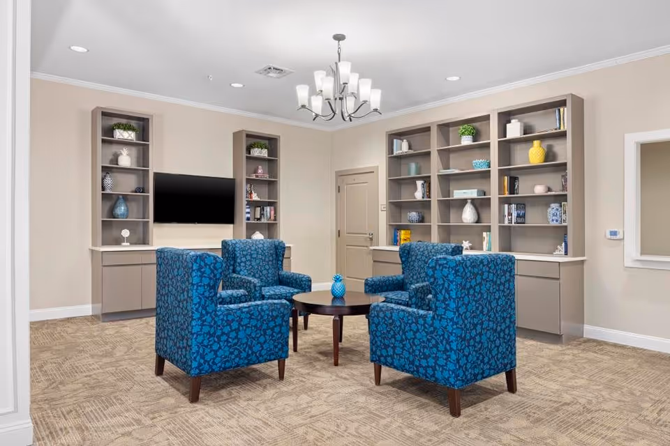 A cozy sitting area in a senior living facility featuring four blue patterned armchairs arranged around a round wooden coffee table with a small decorative blue vase. Behind the chairs are built-in shelves with various decorative items and books, and a flat-screen TV mounted on the wall. The room has beige walls, carpeted flooring, and a modern chandelier hanging from the ceiling.