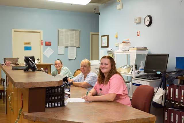 Three female staff members sitting behind a reception desk in a healthcare facility, smiling towards the camera. The room has light blue walls, office equipment, and paperwork visible in the background.