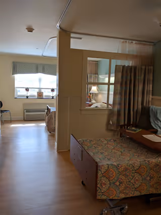Interior view of a nursing and rehabilitation center room with a hospital bed covered in a patterned blanket, a privacy curtain partially drawn, a small table with items on it, and a window with blinds and valance in the background letting in natural light.