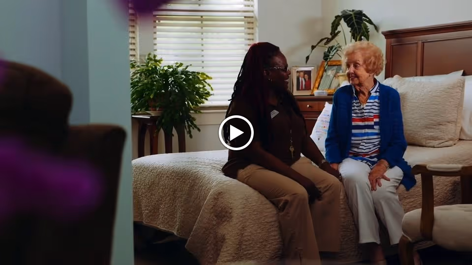 An elderly woman and a caregiver sitting on the edge of a bed in a bedroom, engaged in conversation. The room has a window with blinds, a plant on a stand, framed photos on a dresser, and a cushioned chair nearby.