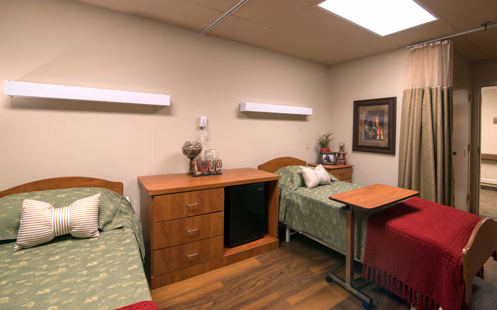 A cozy room with two single beds, each with green patterned bedding and a decorative pillow. Between the beds is a wooden cabinet with drawers and a small black refrigerator. A red throw blanket is draped over the foot of the right bed, which also has an adjustable overbed table. The walls are beige, with two horizontal wall lights above the beds and a framed picture on the right wall. A curtain partially covers a doorway in the background.