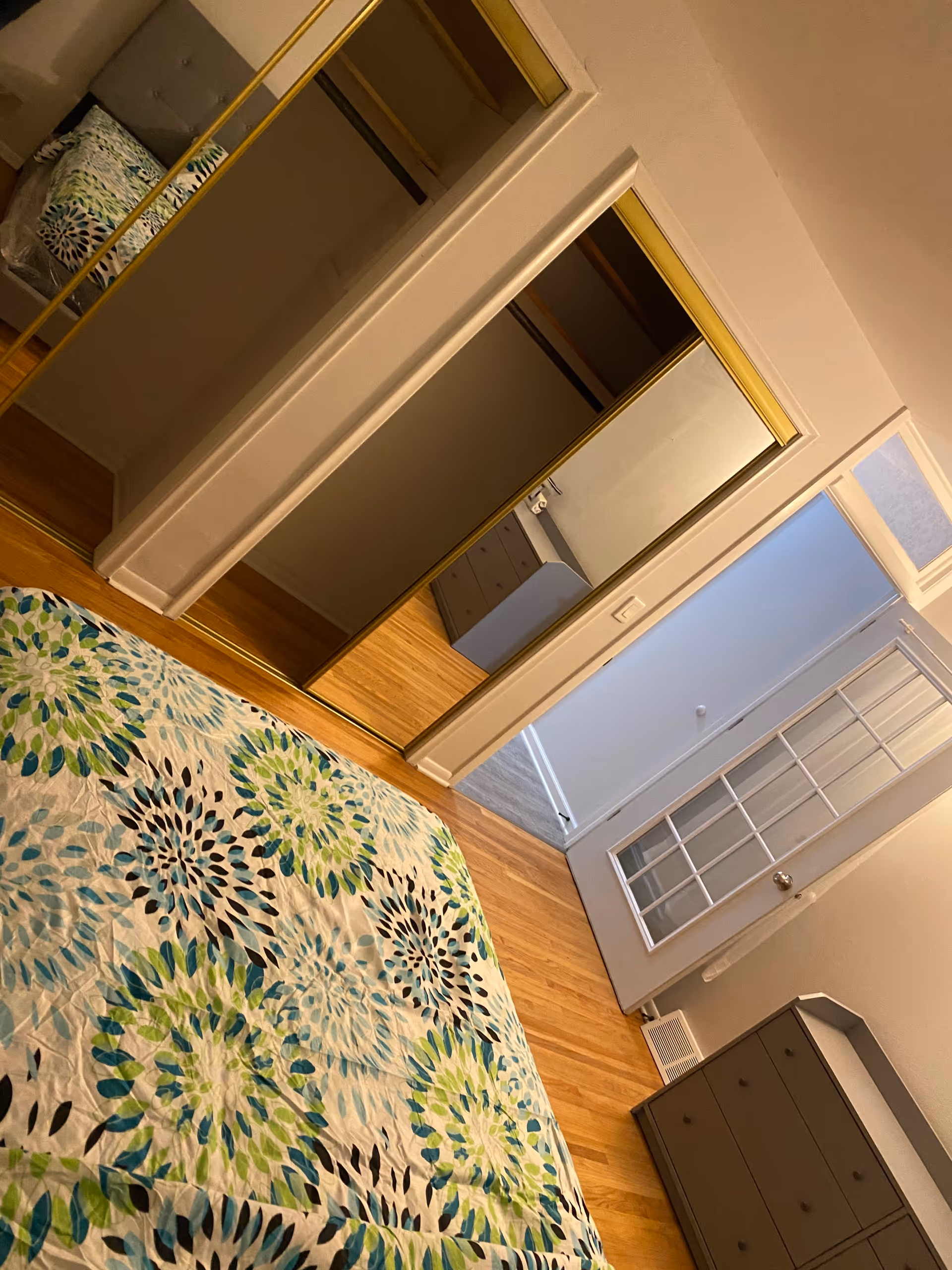 A bedroom with a bed covered in a floral patterned bedspread in shades of green, blue, and black. There is a mirrored sliding closet door with a gold frame, a gray dresser, and a white door with glass panels leading to another room. The floor is wooden and the walls are painted white.