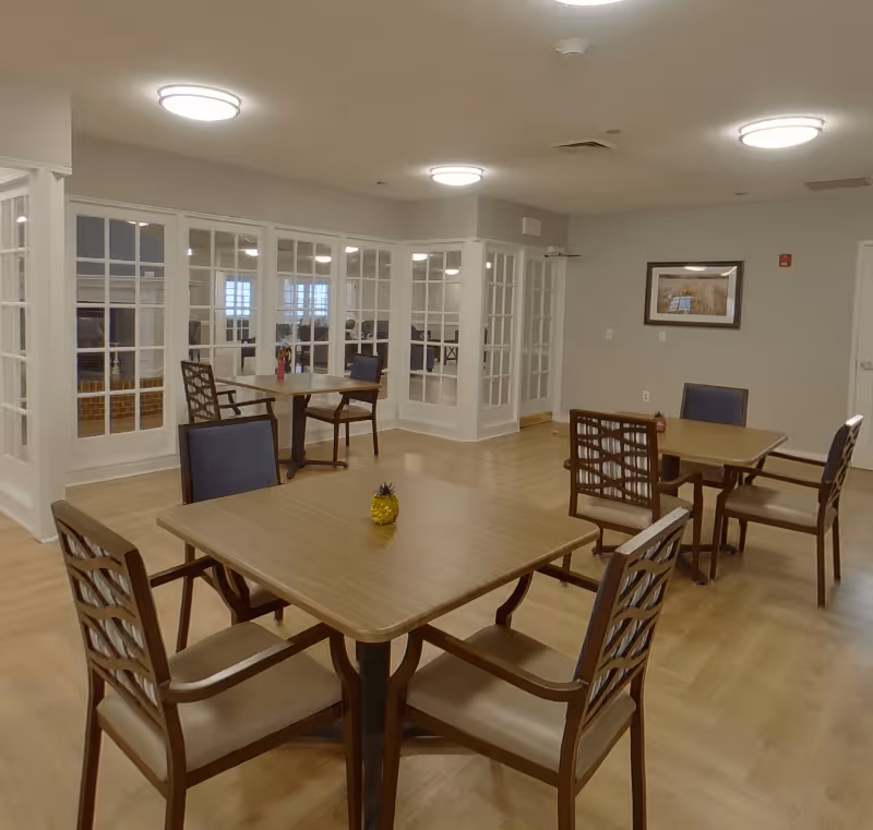 A well-lit dining area with wooden tables and chairs arranged neatly on a light wood floor. The room features large glass-paneled walls and doors, with a framed picture hanging on the far wall. The ceiling has round light fixtures providing ample illumination.