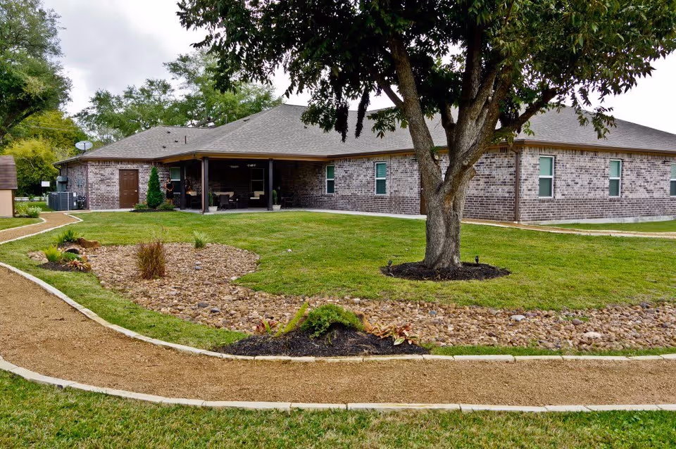 A single-story brick building with a covered patio area, surrounded by a well-maintained lawn, a large tree, and landscaped garden beds with rocks and plants. A gravel pathway curves through the grassy area in front of the building.