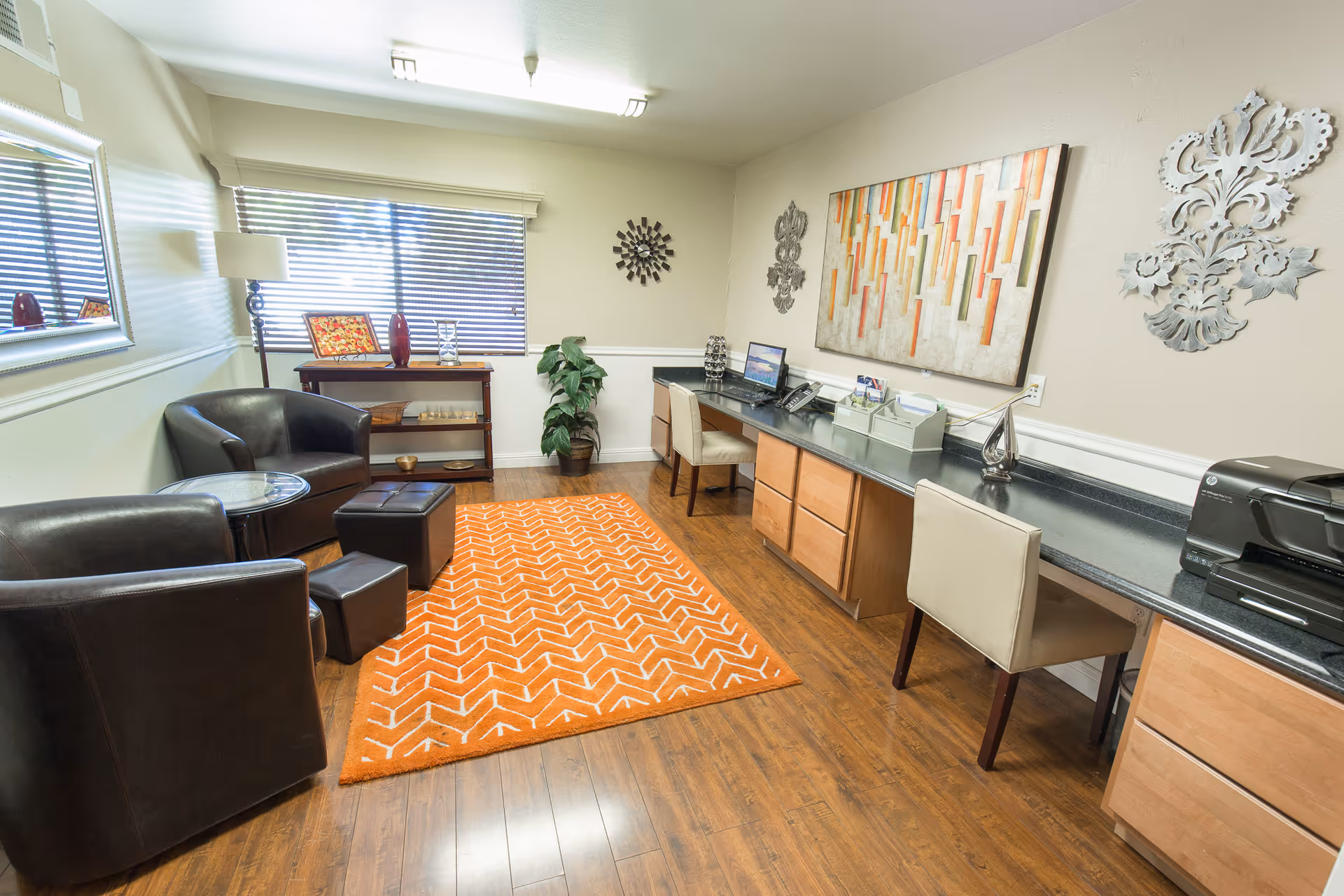 Bright communal office lounge with desk workstations along one wall, a seating area of leather chairs and ottomans, and an orange patterned rug on wood floors.