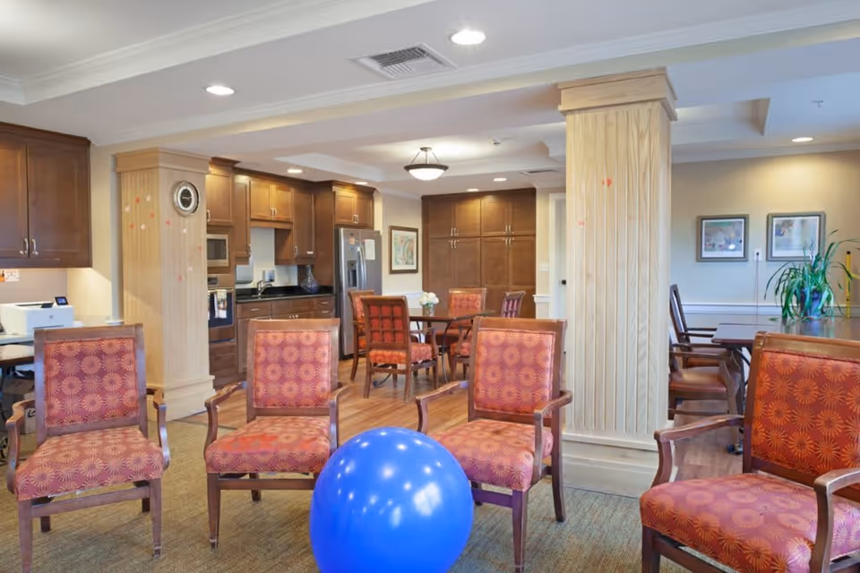 A spacious and well-lit common area with wooden flooring and multiple wooden chairs upholstered in red patterned fabric arranged around a blue exercise ball. The background features a kitchen area with wooden cabinets, a refrigerator, and a microwave. There are two large wooden pillars, framed artwork on the walls, and a plant on a table to the right.