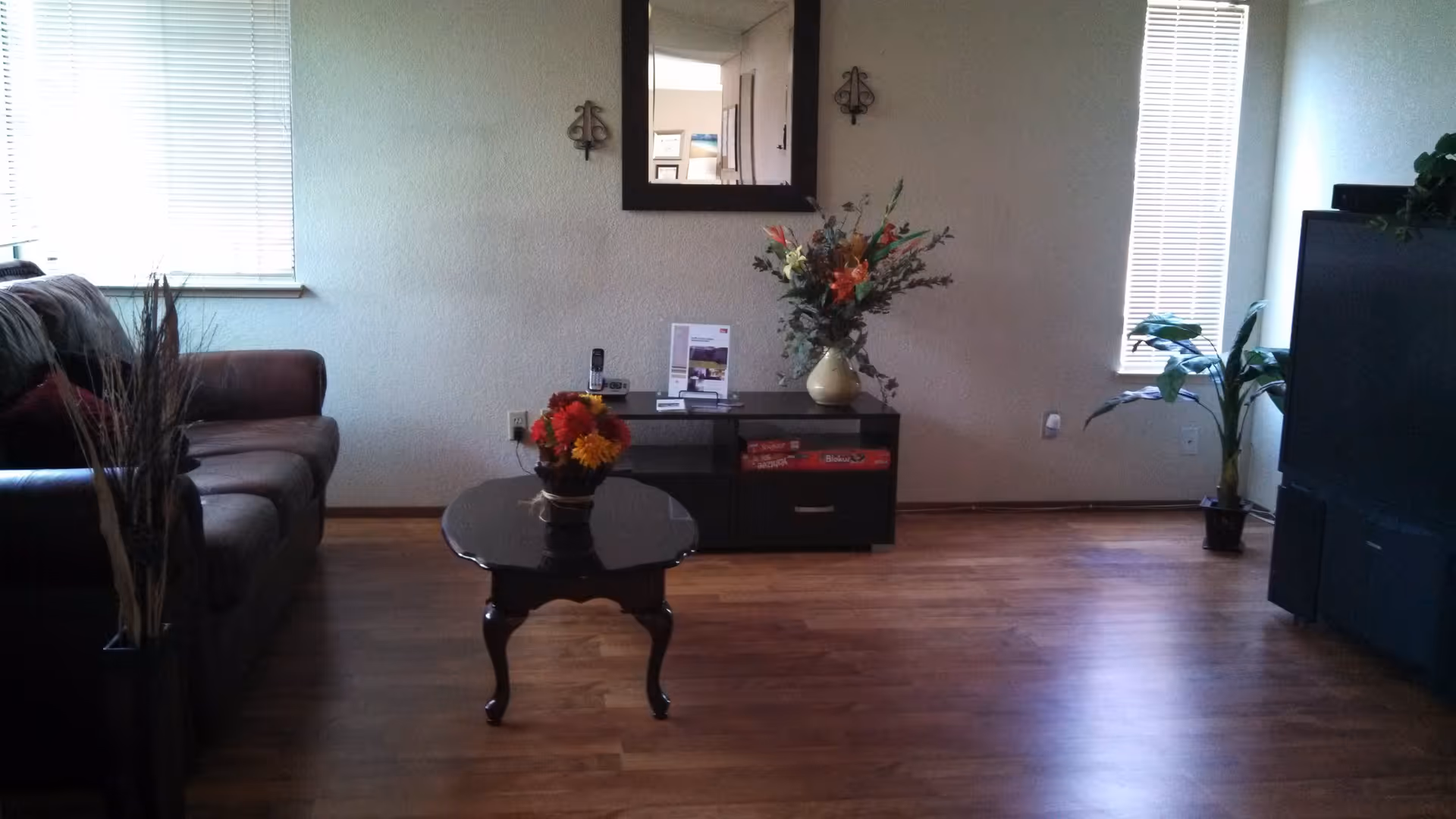 A living room with a dark brown sofa on the left, a small dark wooden coffee table with a flower arrangement in the center, and a black TV stand with a large TV on the right. There is a mirror on the wall above the TV stand, flanked by two decorative wall sconces. The room has wooden flooring and two narrow windows with blinds, one on each side of the TV stand. A potted plant is placed near the TV stand.