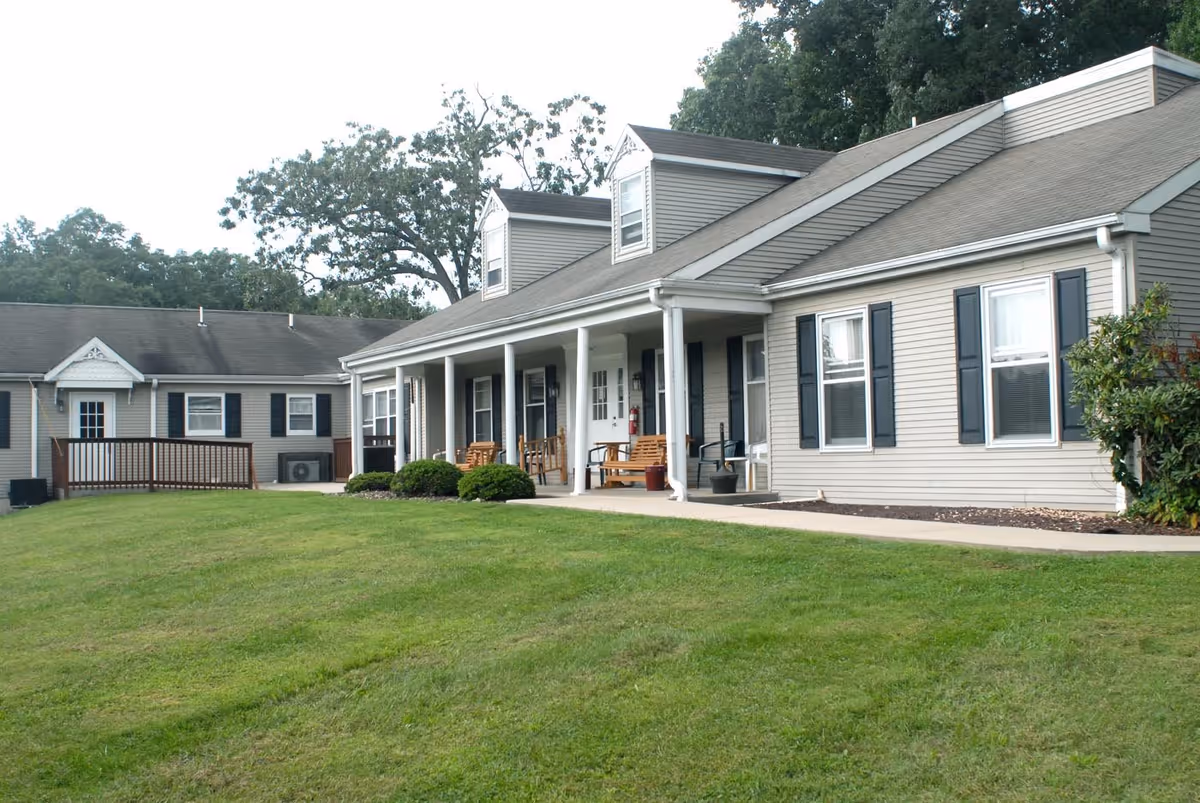 Exterior view of a single-story residential building with beige siding, black shutters, and a covered porch with wooden benches. The building is surrounded by a well-maintained green lawn and some bushes, with trees in the background.