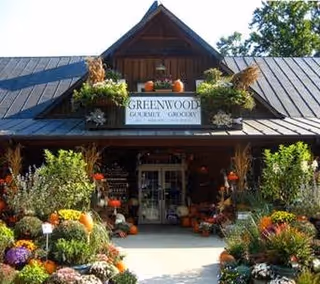 Exterior view of a rustic wooden building with a peaked roof, featuring a sign that reads 'Greenwood Gourmet Grocery'. The entrance is surrounded by abundant colorful flowers, plants, and pumpkins, creating a welcoming and vibrant atmosphere.