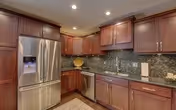 Modern kitchen with stainless steel refrigerator, dark wood cabinets, granite countertops, and a tiled backsplash. The kitchen is well-lit with recessed ceiling lights.