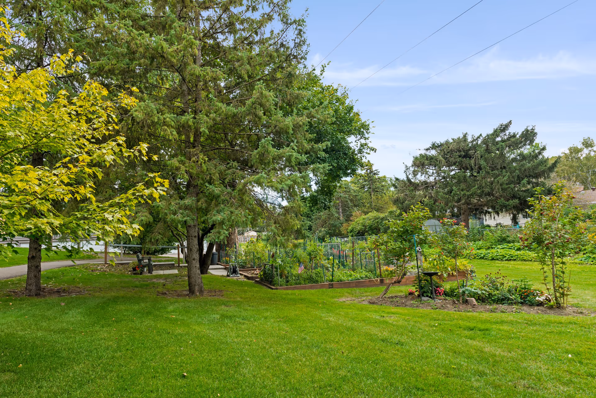 A lush green outdoor garden area with various trees, plants, and a small fenced vegetable or flower garden. The sky is clear with a few clouds, and there is a pathway visible on the left side.