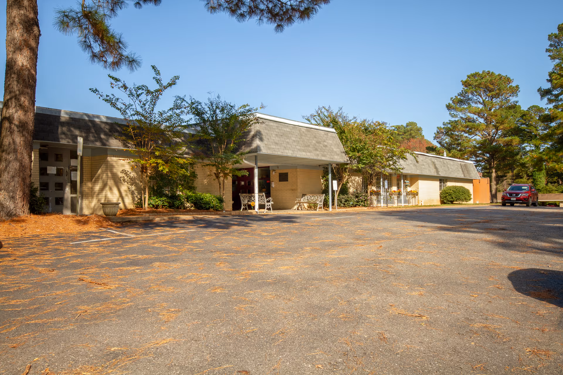 Exterior view of The Blossoms at Stamps Rehab & Nursing Center showing a single-story building with a gray roof and beige brick walls. There are trees and shrubs around the building, a few benches near the entrance, and a parking lot with a red car parked on the right side. The sky is clear and blue.
