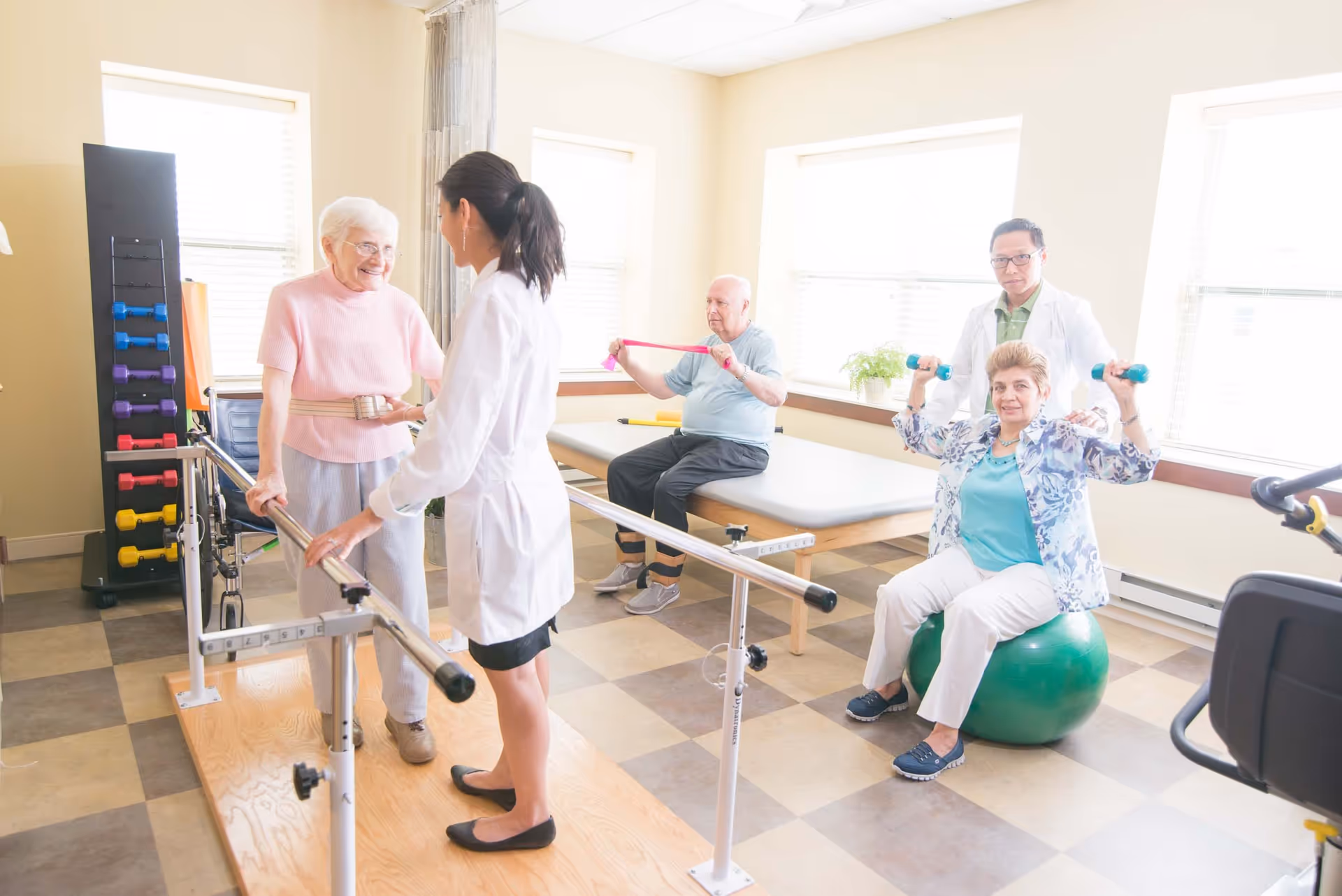 A senior living facility physical therapy room where an elderly woman is walking with the assistance of a female therapist holding parallel bars. In the background, an elderly man is exercising with a resistance band while seated on a therapy table, and another elderly woman is lifting small dumbbells while sitting on an exercise ball, assisted by a male therapist. The room is well-lit with large windows and has various exercise equipment including dumbbells and a wheelchair.