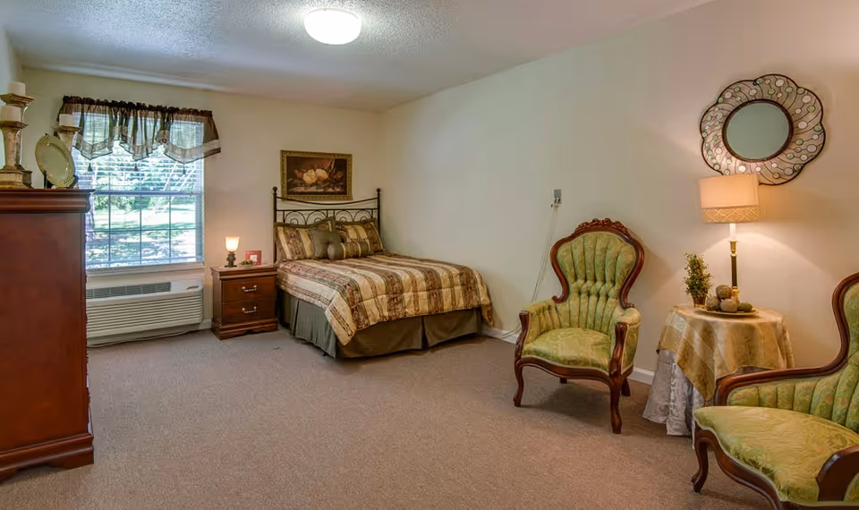 A cozy bedroom in a senior living facility with a single bed covered in a patterned quilt, a wooden nightstand with a lamp, two vintage green upholstered armchairs, a small round table with a lamp and decorative items, a large window with sheer curtains, and a wooden dresser. The walls are light-colored and there is a decorative mirror and framed artwork on the walls.