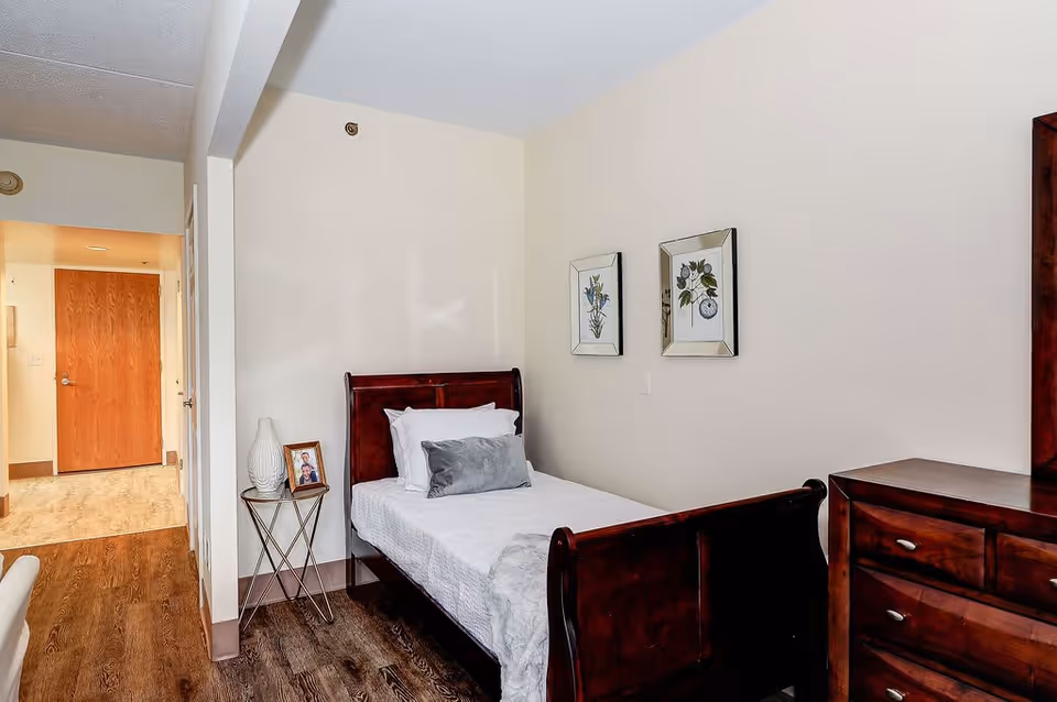 A small bedroom with a single dark wooden bed made with white bedding and a gray pillow. Next to the bed is a small round side table with a white vase and a framed photo. Two framed botanical prints hang on the wall above the bed. A dark wooden dresser is partially visible on the right side. The room has light-colored walls and wood flooring, with a hallway and a closed wooden door visible in the background.