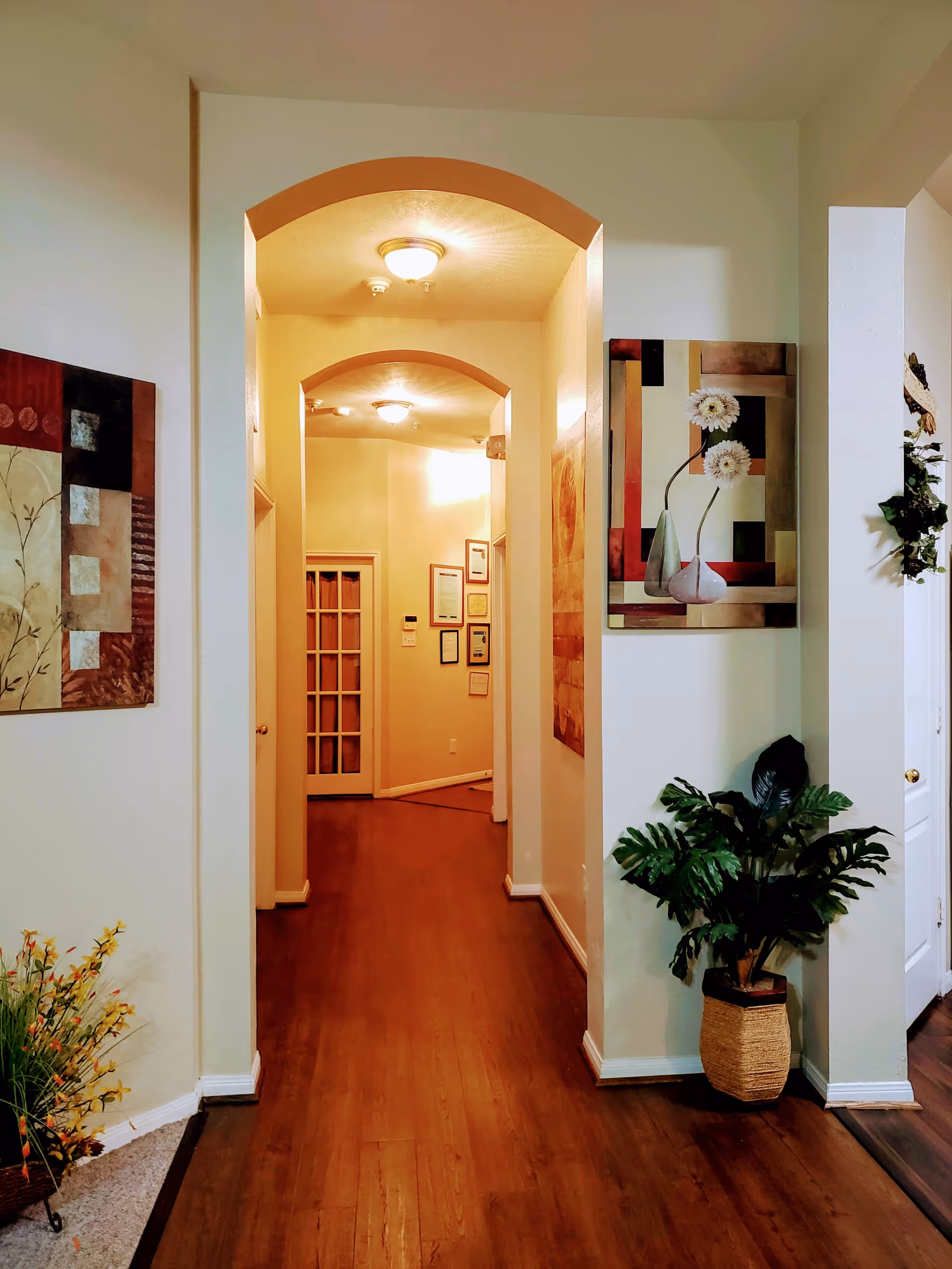 Interior hallway of a senior living facility with wooden flooring, cream-colored walls, and arched doorways. The hallway is decorated with framed artwork on the walls and a potted plant on the right side. There are ceiling lights illuminating the space and a door with glass panels at the end of the hallway.