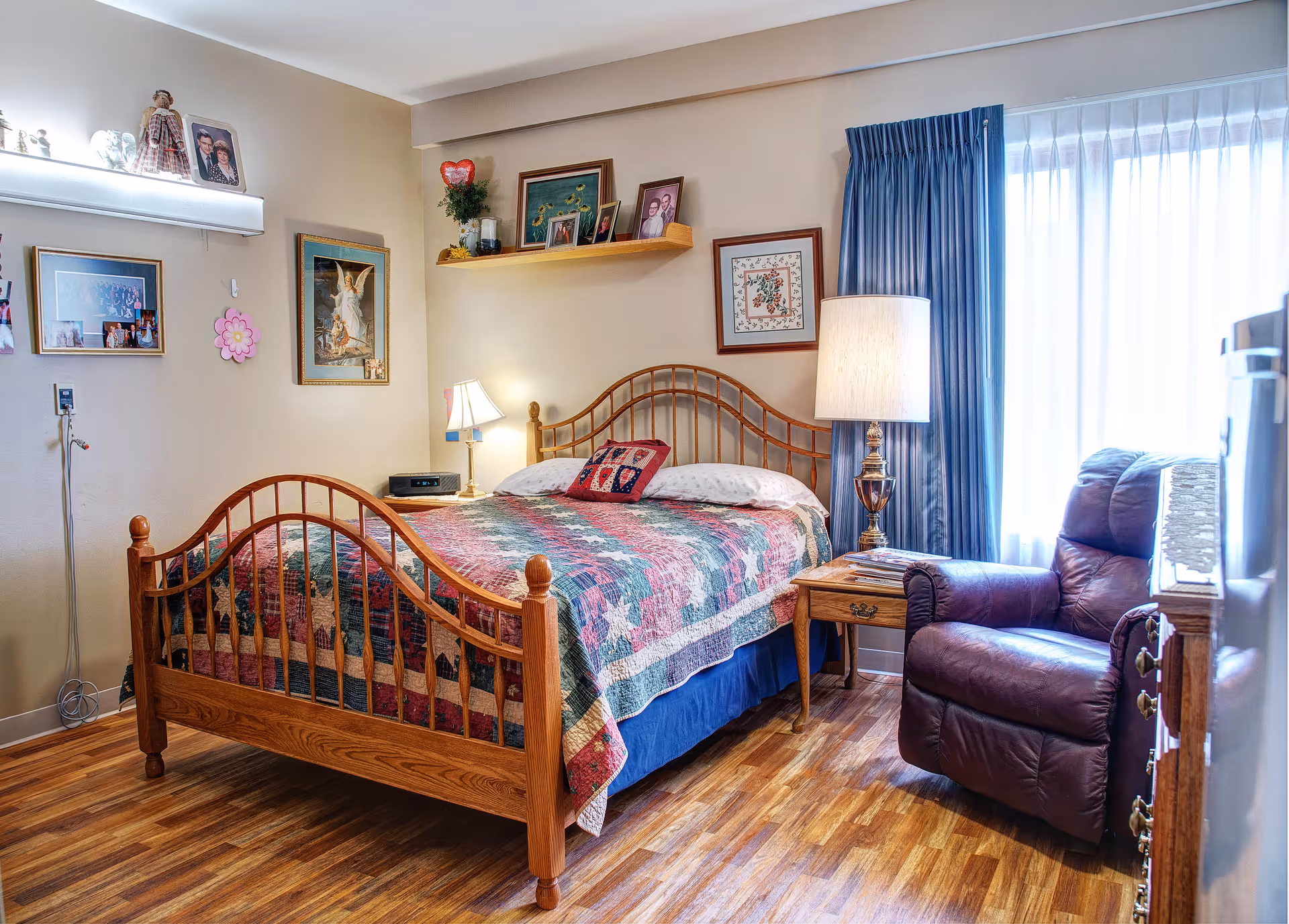 Cozy senior bedroom with a wooden bed dressed in a patchwork quilt, bedside tables and lamps, a recliner by a curtained window, and framed wall decor.