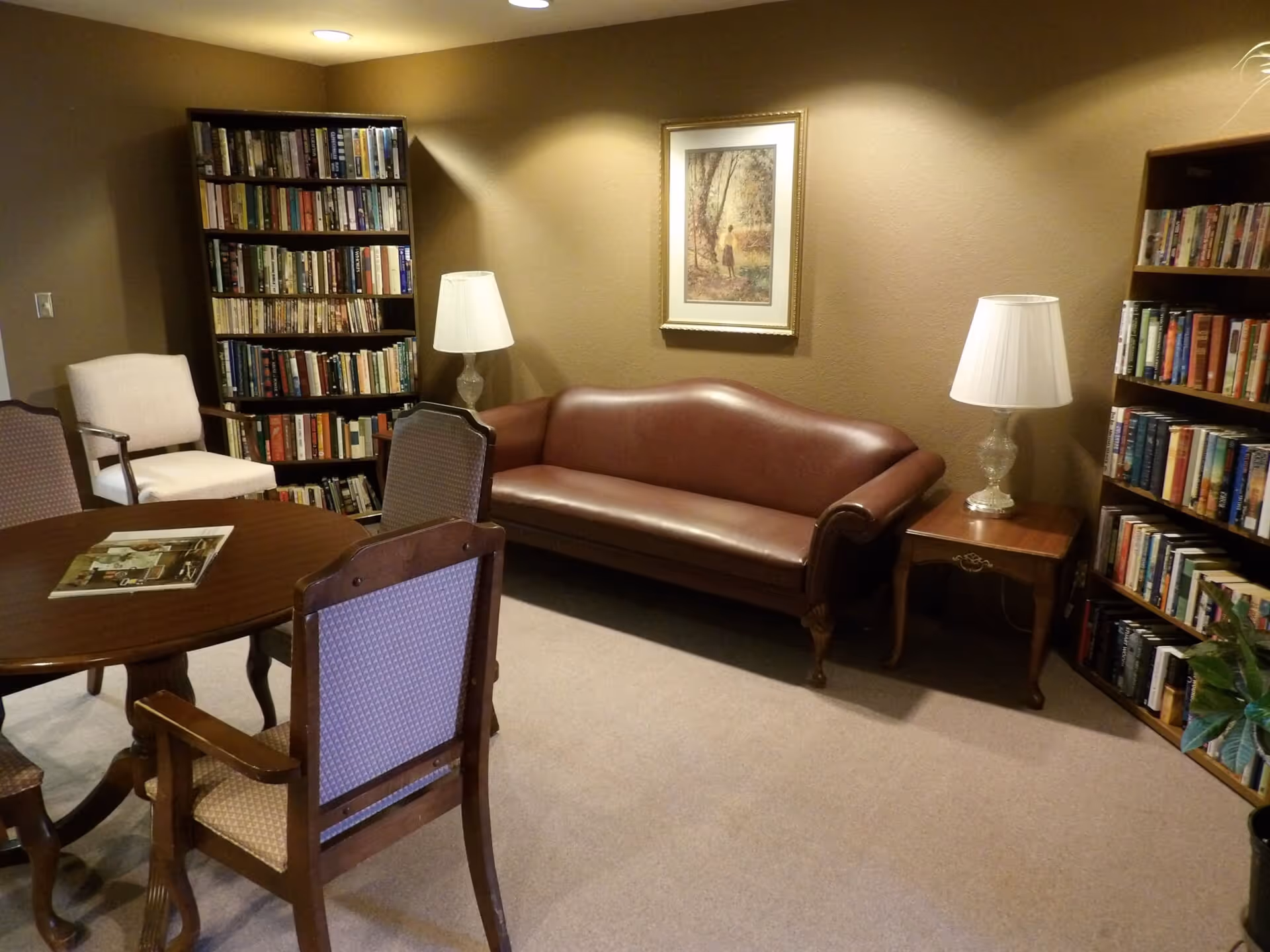 A cozy reading or sitting room with a brown leather sofa, two wooden bookshelves filled with books, a round wooden table with four chairs, two table lamps with white shades on wooden side tables, and a framed painting on the beige wall.