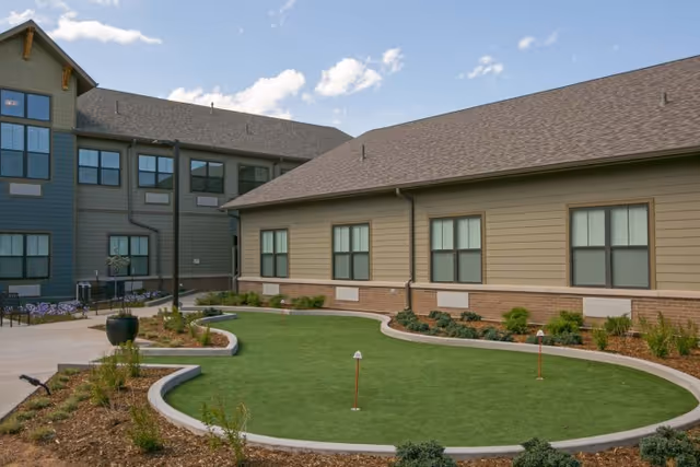 Outdoor courtyard area with a putting green surrounded by a curved concrete border, landscaping with plants and shrubs, and two buildings with multiple windows under a partly cloudy sky.