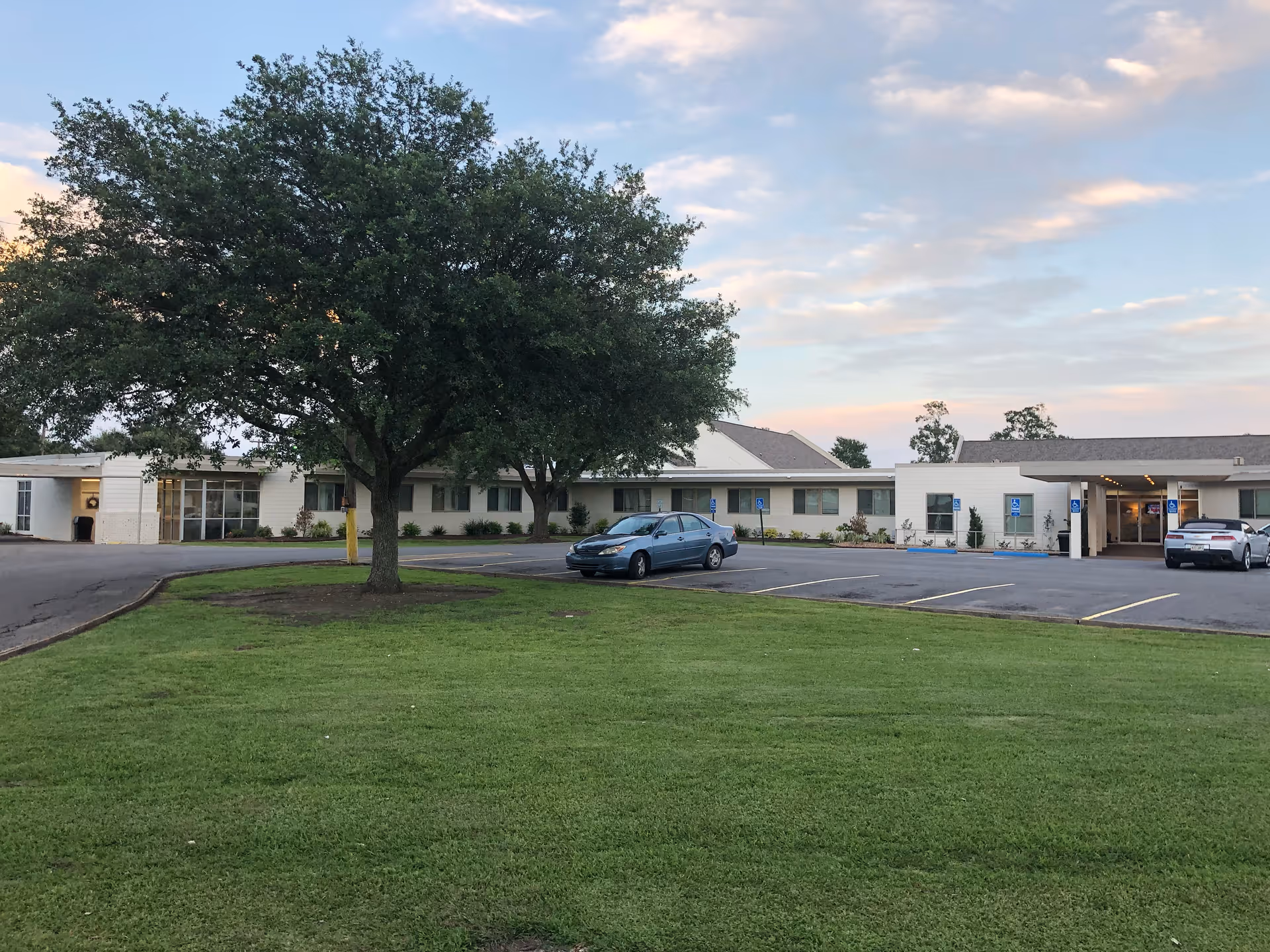 Exterior view of a single-story senior living facility building with a large tree and green lawn in the foreground, a parking lot with a few cars, and a partly cloudy sky at sunset.