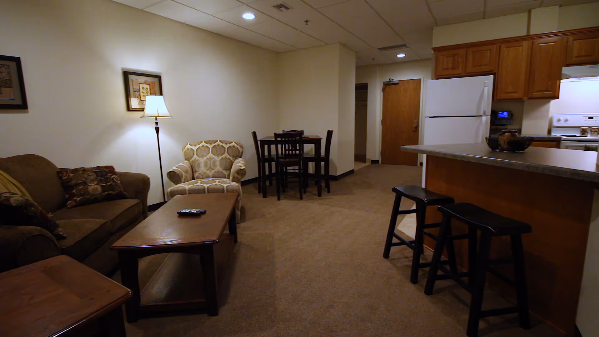 Interior view of a living space in Sherwood Lodge Assisted Living featuring a brown sofa with patterned cushions, a beige armchair, a wooden coffee table with a remote control, a floor lamp, a small dining table with four chairs, and a kitchen area with a refrigerator, stove, and two black bar stools at a counter.
