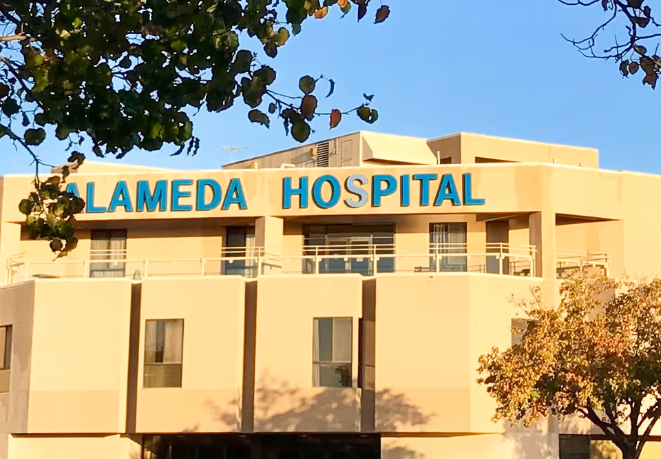 Exterior view of Alameda Hospital building with beige walls and blue signage reading 'ALAMEDA HOSPITAL' under a clear blue sky, partially framed by tree branches with green and brown leaves.