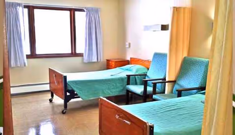 Two hospital beds with green sheets, blue chairs, and a window in a patient bedroom.