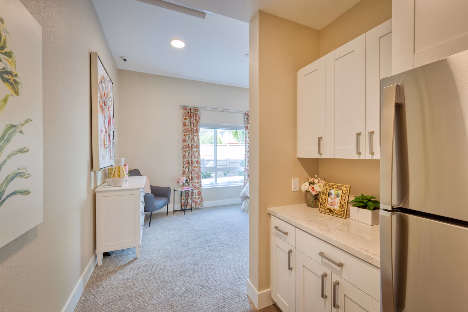 Interior view of a senior living facility room at Ivy Park at San Tomas showing a small kitchenette area with white cabinets, a stainless steel refrigerator, and a countertop decorated with a framed photo, flowers, and a small plant. Adjacent to the kitchenette is a carpeted living space with a white dresser, a gray armchair, a small side table with flowers, a large window with floral curtains, and light beige walls.