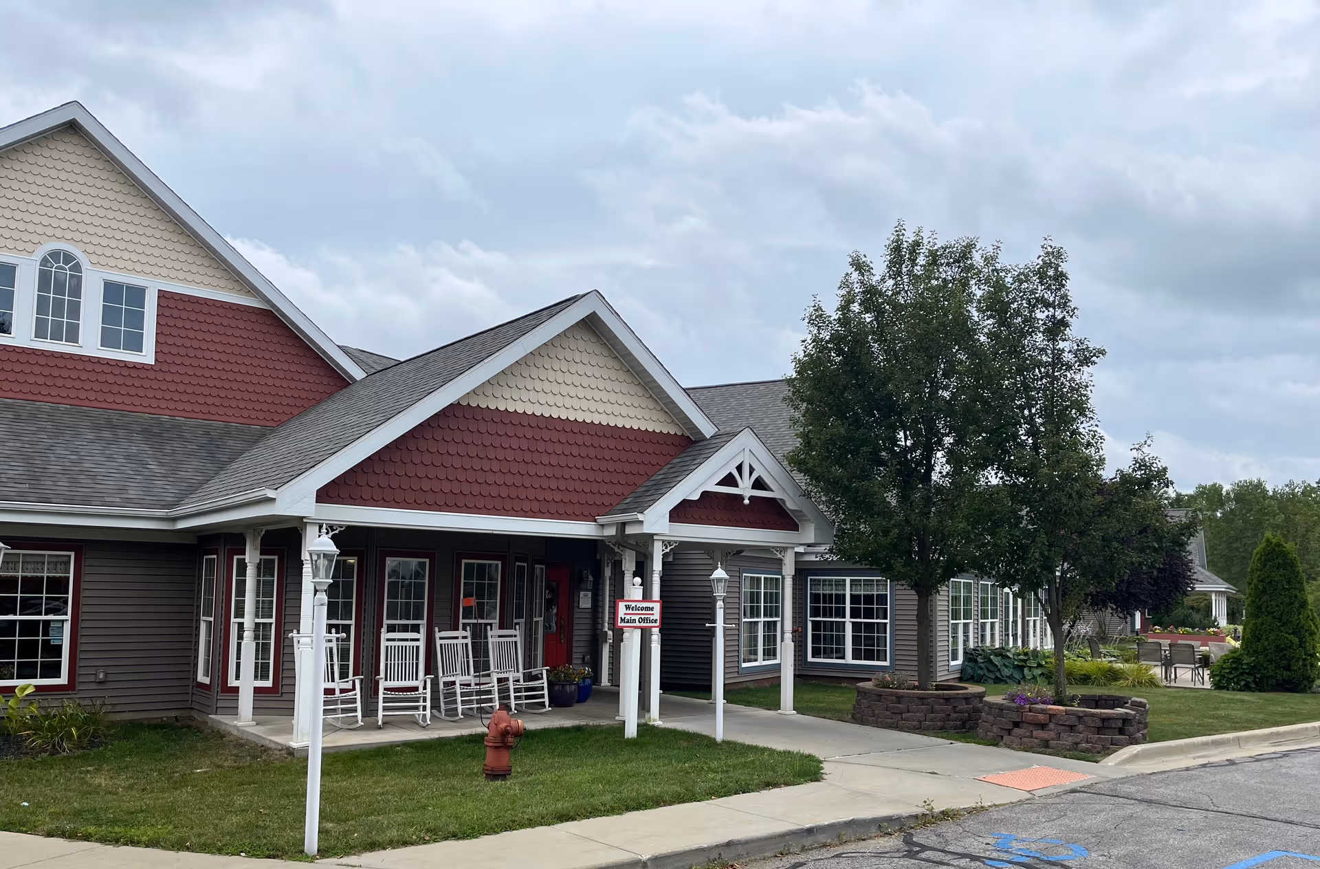 Exterior view of Caretel Inns of St. Joseph building with a covered entrance, white rocking chairs on the porch, a red fire hydrant on the grass, and a sign that reads 'Welcome Main Office'. There are trees and landscaped areas around the building under a cloudy sky.