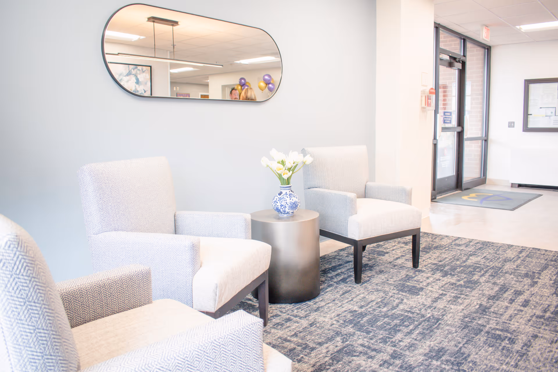 A bright lobby seating area with upholstered chairs around a small table holding a vase of flowers, a wall mirror, and glass entry doors in the background.