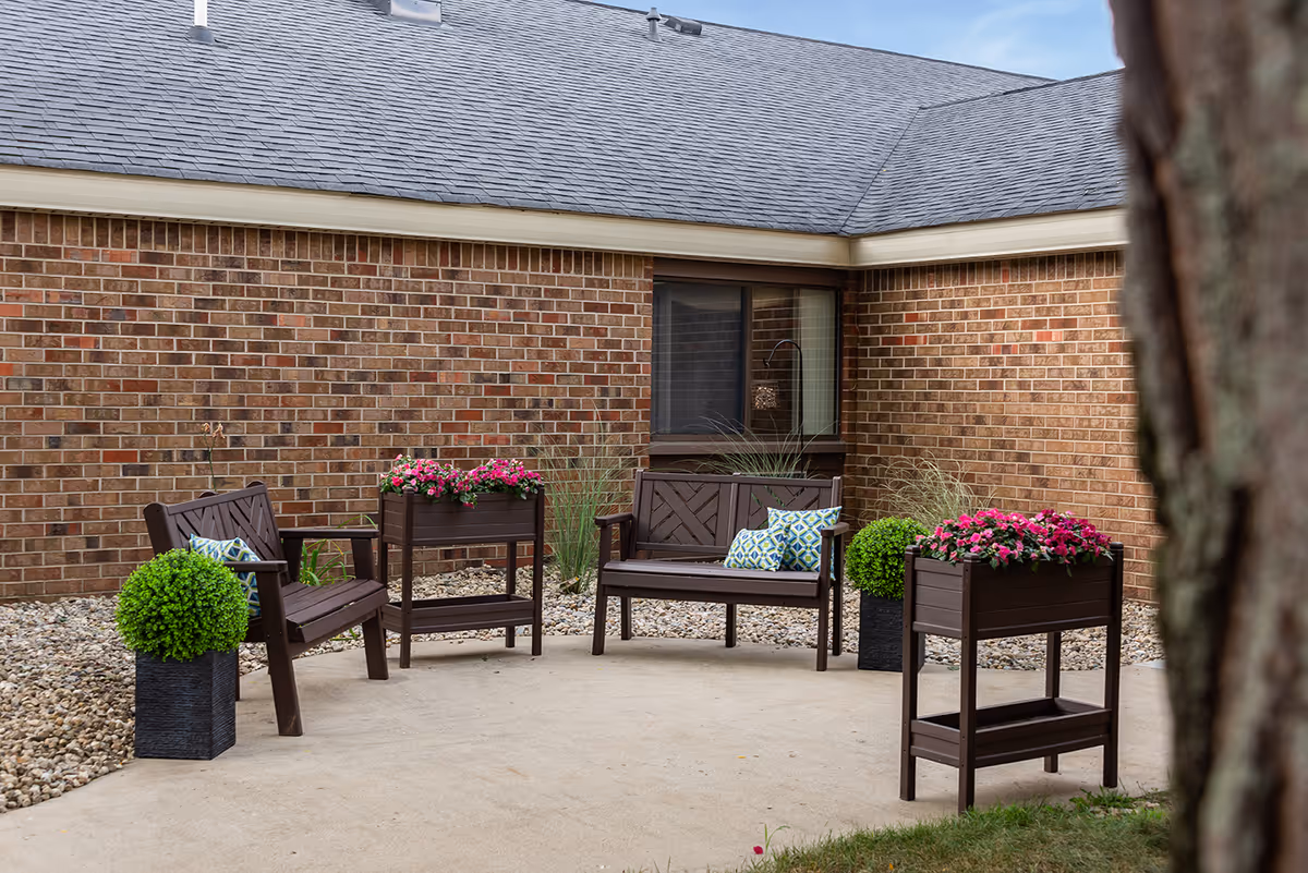 Outdoor patio area with two wooden benches and two wooden chairs, each with decorative cushions. There are two planters with pink flowers and two planters with green shrubs on a concrete surface, surrounded by a brick building and gravel landscaping.