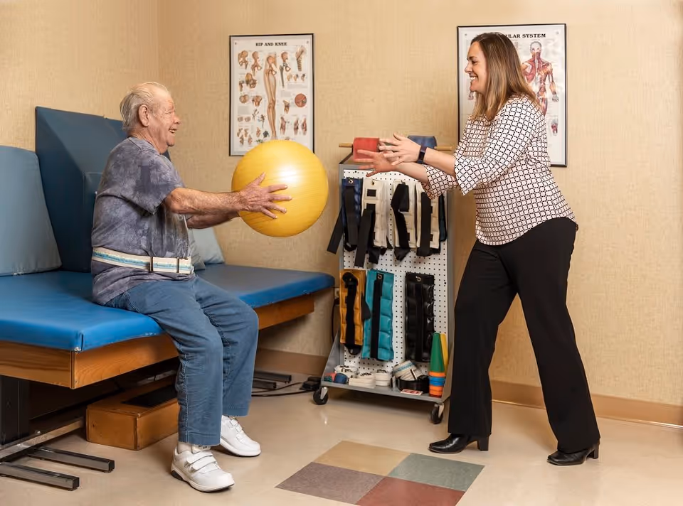 An elderly man sitting on a padded examination table in a rehabilitation room, passing a yellow exercise ball to a smiling female therapist standing in front of him. The room has anatomical posters on the wall and a rack with various therapy equipment.