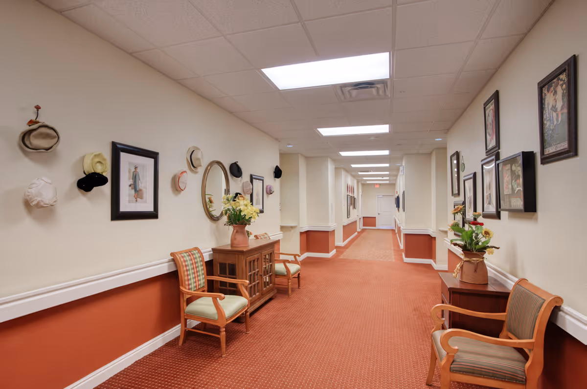 Carpeted hallway in a senior living facility with chairs, framed artwork, decorative hats on the walls, and small tables with flower arrangements.
